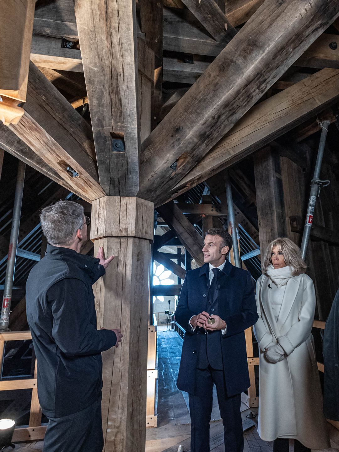 Brigitte, Emmanuel, and officials stand beneath intricate wooden beams of a reconstructed cathedral spire. Brigitte’s white coat contrasts with the rustic setting as a guide explains the structure.