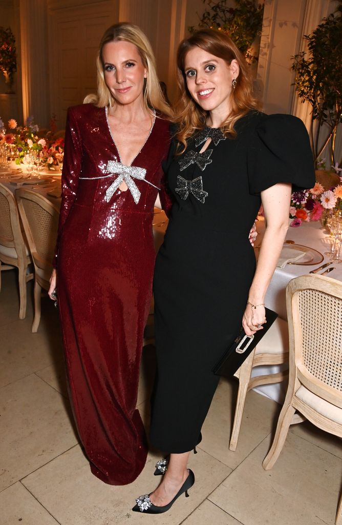 Princess Beatrice in a black dress with sparkly bows at the front posing with a friend in a burgundy sequin dress