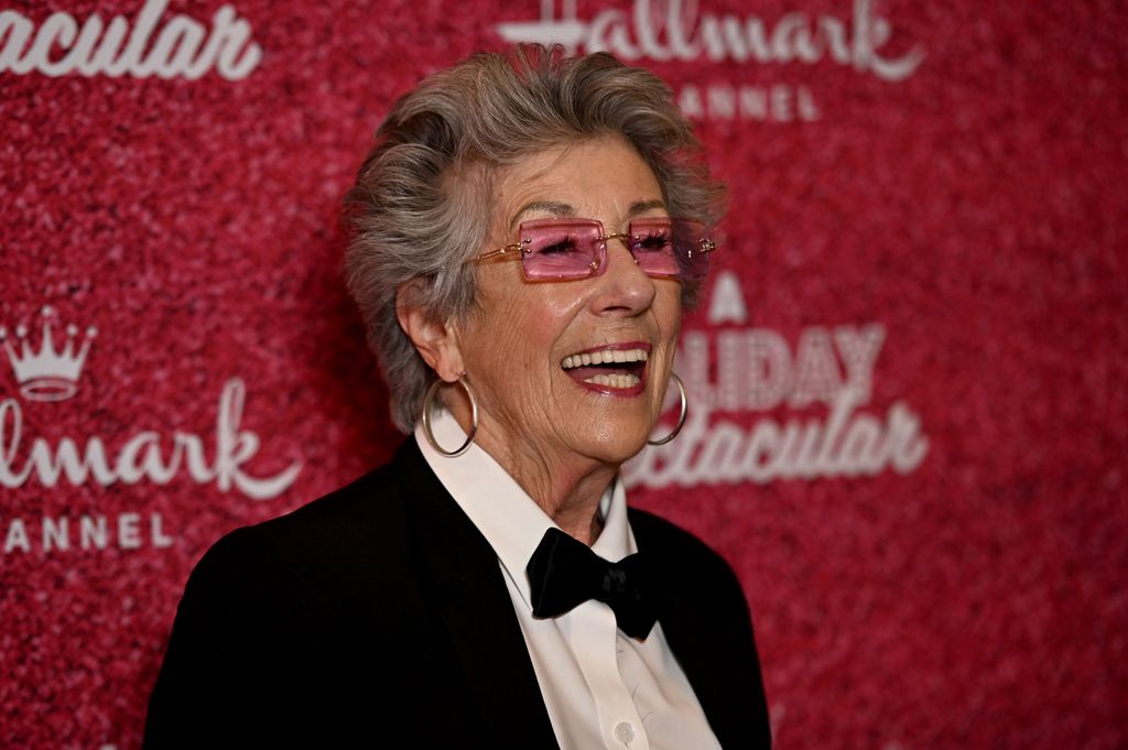 Ellen Travolta smiling in a suit and bowtie against a red background