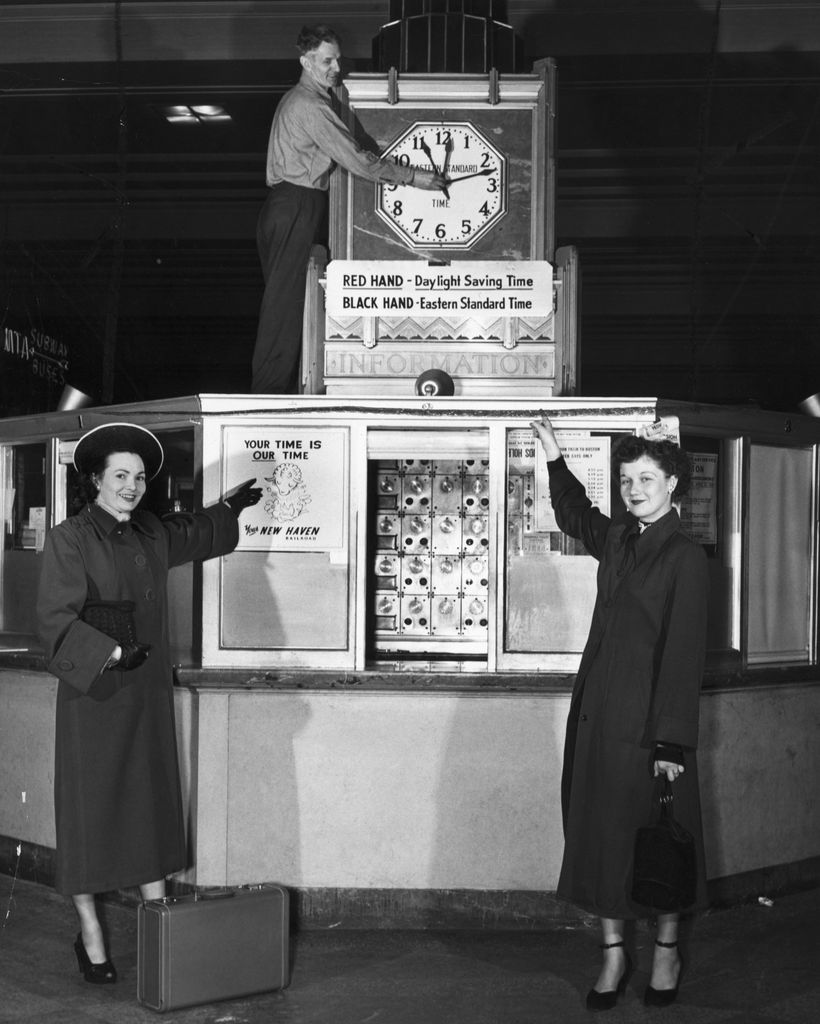 The Misses Margaret L. Dowling (l) and Jean Baker, of Roslindale, get a preview of how the clocks in South Station at Boston will appear after 2 a.m., next Sun. morning after all of us set our watches and clocks ahead one hour to conform to Daylight Saving Time. Electrical foreman Frank L. Wilson tests out the extra red hou-hand which will appear on all the South Station clocks