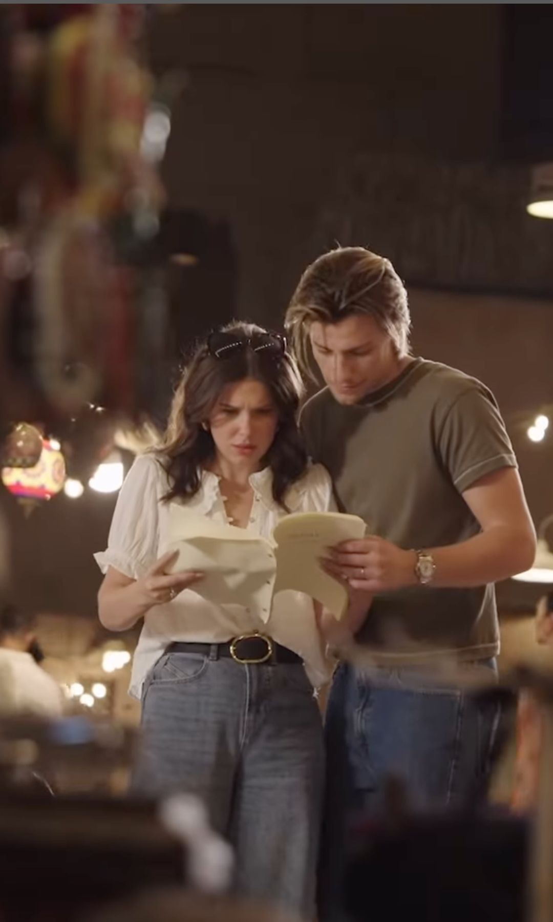 Millie bobby Brown and Jake Bongiovi pore over a script during a BTS moment from their short film about Dubai. Millie wears a white blouse and jeans. Jake wears a khaki tshirt and jeans. 