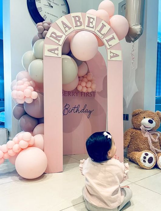 A young girl sat in front of a pink archway and a teddy bear