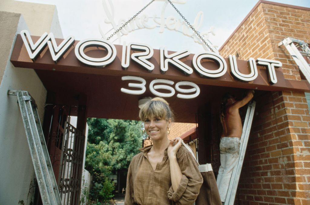 Jane Fonda, wearing a brown checked blouse with the sleeves rolled up, outside her 'Workout' aerobics studio as a workman stands on a ladder to put the finishing touches to the sign before the studio's opening, at 369 South Robertson Boulevard in Beverly Hills, California, 13th September 1979