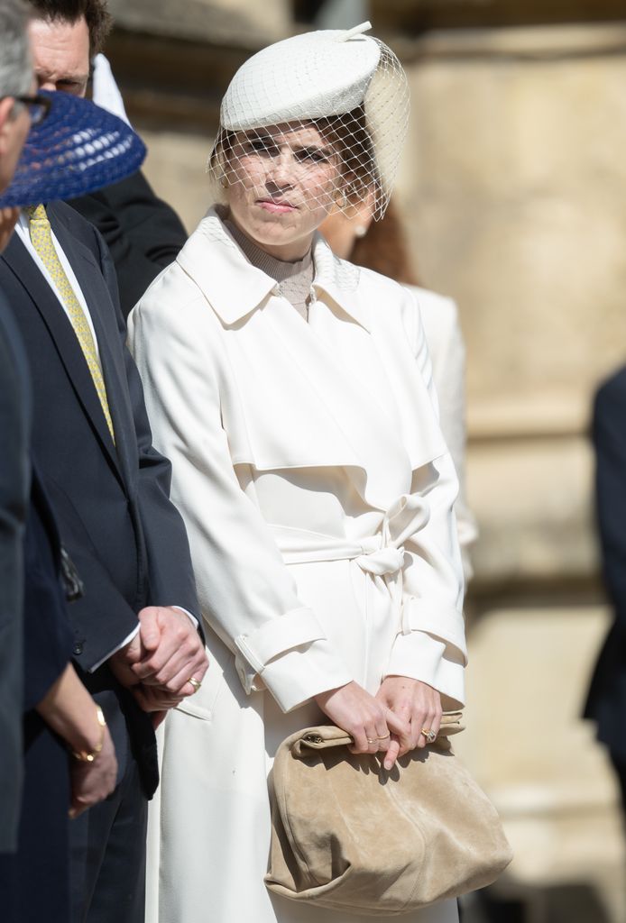 Princess Eugenie in a cream trench coat with a netted hat, holding a tan clutch while standing beside fellow royals outside Windsor’s St George’s Chapel.