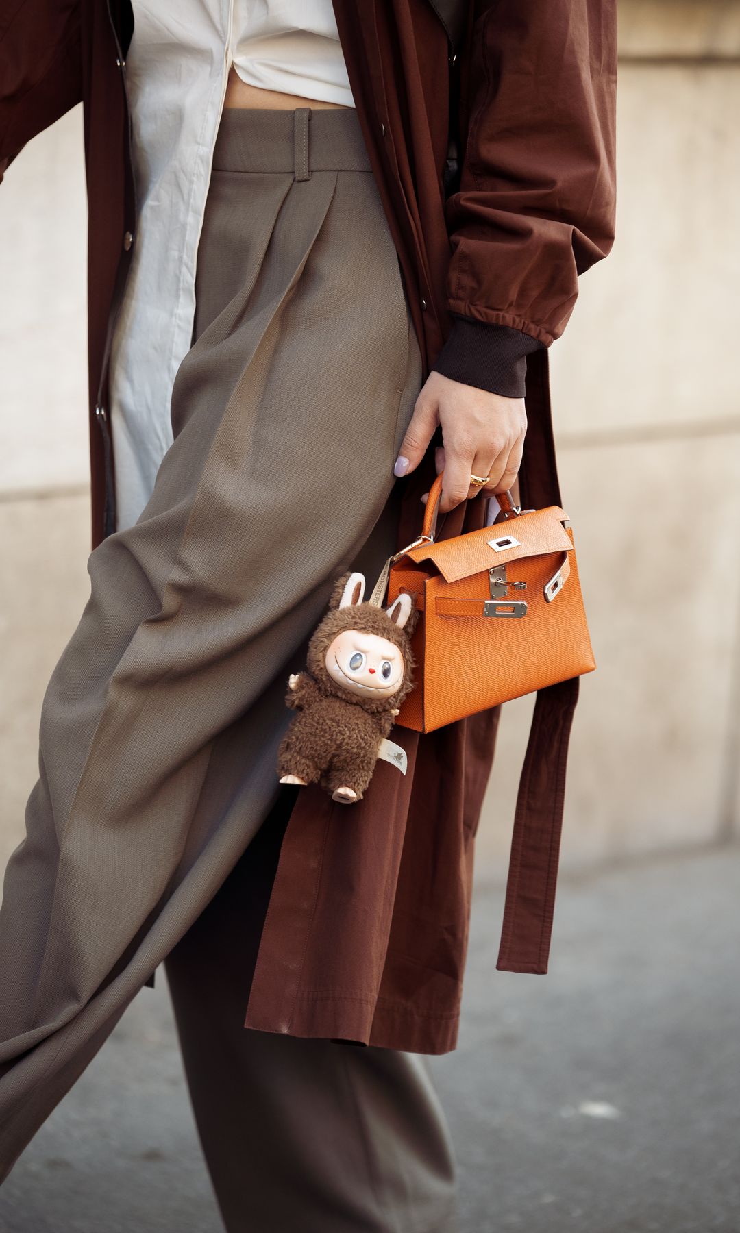 A guest wears brown coat, white blouse, orange Hermes bag with a bag charm brown Labubu monster, grey pants outside the Hermes fashion show during the Womenswear Fall/Winter 2025/2026 as part of Paris Fashion Week on March 08, 2025 in Paris