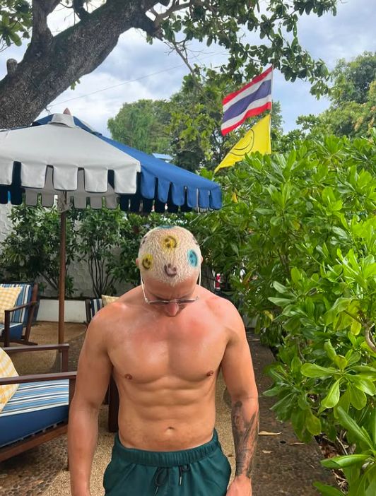A shirtless man standing on a beach; smiley faces have been drawn in his blonde hair
