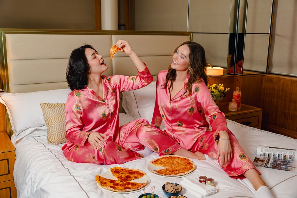 Women wearing silk Their Nibs pyjamas on hotel bed with pizza