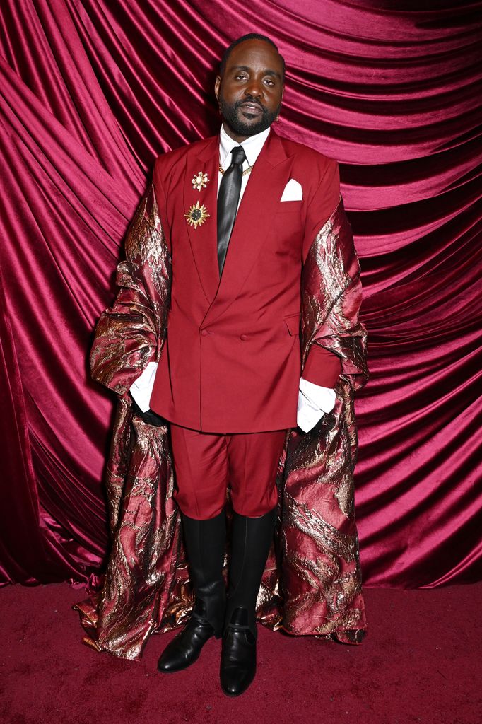 Brian Tyree Henry wears a burgundy short suit with a gold-lined opera cape and knee-high boots at the 2025 Met Gala after-party.