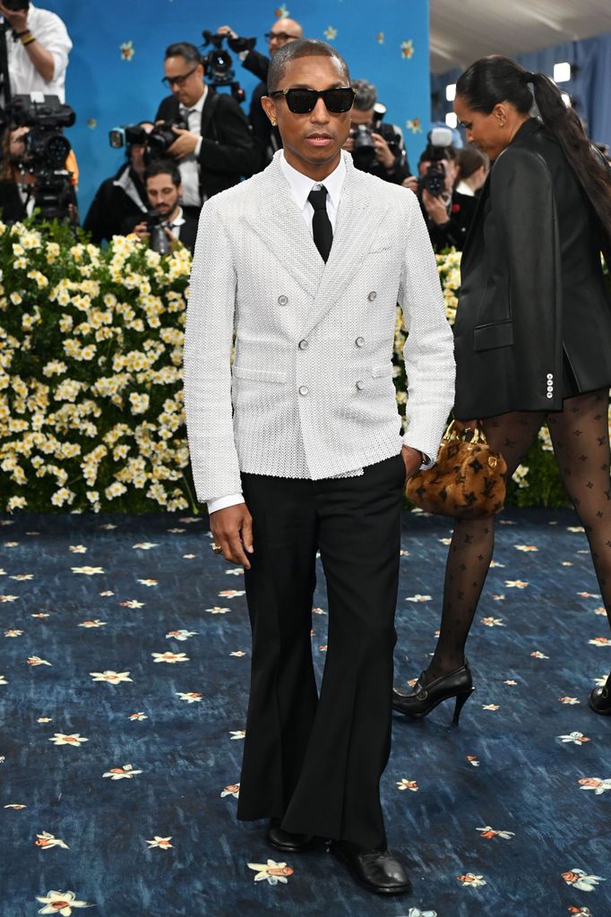 Pharrell Wiliams wears a white double breasted suit jacket, teamed with white shirt and tie and black flared trousers