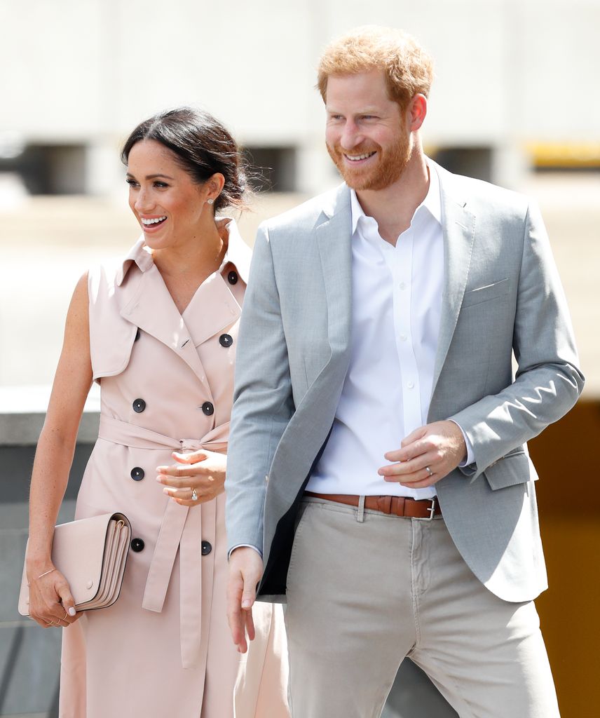 Meghan in pink trench dress and Harry in grey suit smiling