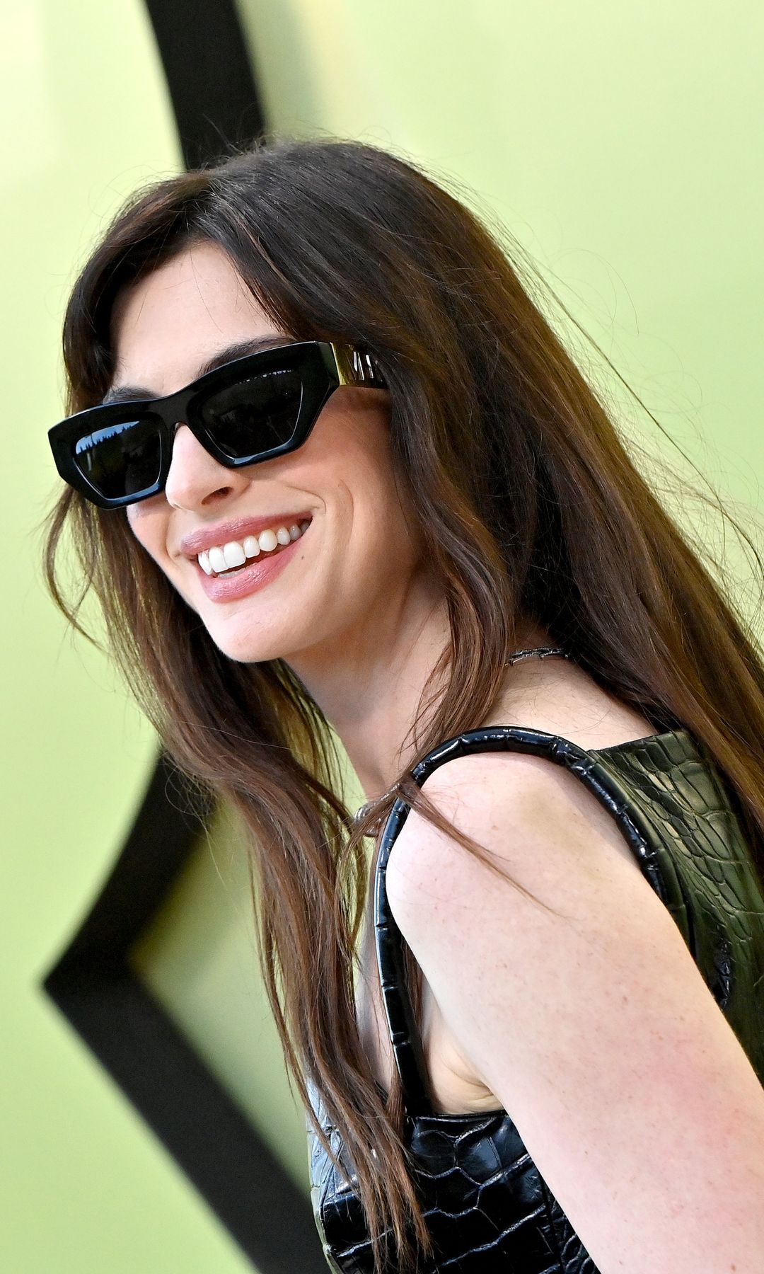 Anne Hathaway is standing against a soft chartreuse and pale lime gradient backdrop with oversized, glossy lettering. She wears a short, fitted black mini dress with a high-gloss crocodile texture. Her legs are covered in sheer black tights, and she wears ultra-high black platform boots with metal buckles. A sleek black handbag with a gold clasp dangles from her hand, and she finishes the look with a silver necklace, bracelets, and black sunglasses. Her long brown hair is styled straight with a middle part.