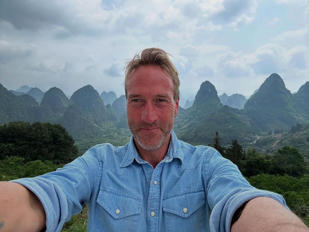 Ben Fogle taking a selfie in front of green mountains