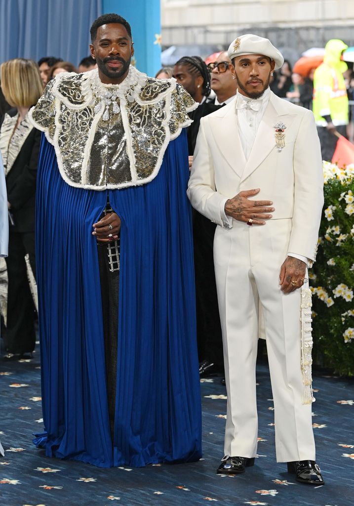 Colman Domingo wears a black cape with intricate silver beaded detail and Lewis Hamilton wears a white suit with cropped jacket, black shoes and a white beret