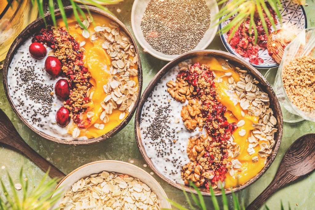 stock image of smoothie mango bowls with chia seeds yogurt pudding and cranberries, nuts, oatmeal topping in coconut shells with spoon, top view