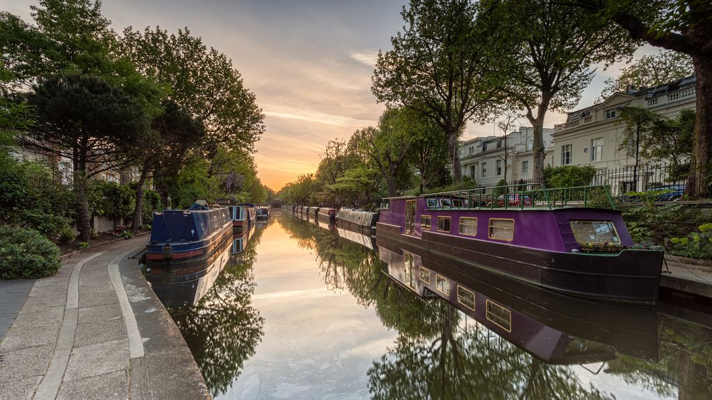 Image shows narrow boats moored on either side of the Regents Canal, London.  The River is calm, still and reflective during the early morning sunrise.