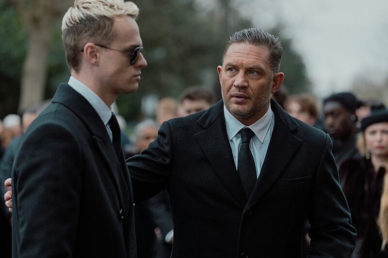 man in black suit escorting younger man through crowd