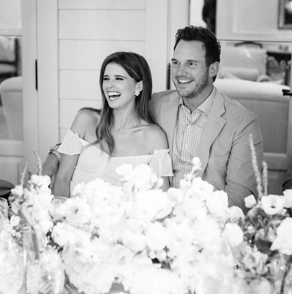 Chris Pratt and Katherine Schwarzenegger sitting together and smiling with flowers in front of them in a black and white photo from their wedding