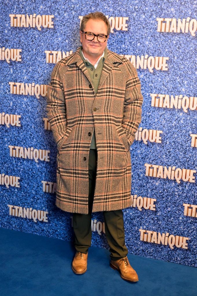 Alan Carr wearing checkered coat standing in front of 'Titanique' step and repeat at the West End opening