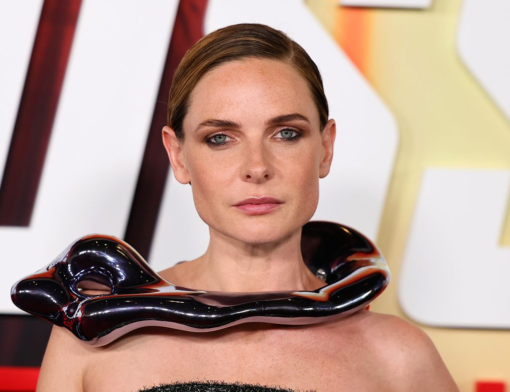 Rebecca wears a sleek metallic sculptural collar accessory with pulled-back hair and smoky makeup, exuding modern elegance at a premiere.