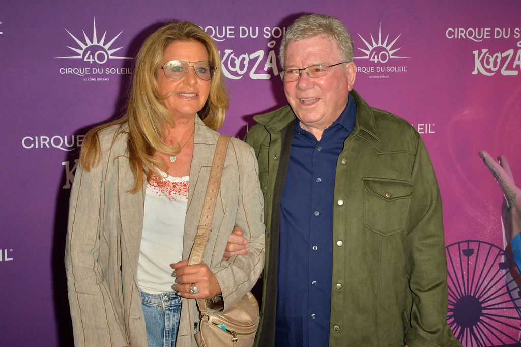 Elizabeth and William Shatner dressed in smart casual attire in front of Cirque Du Soleil event poster smiling for cameras