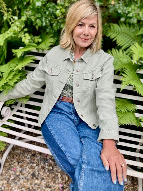 Glynis Barber sitting  on a bench in a green denim jacket and blue denim jeans
