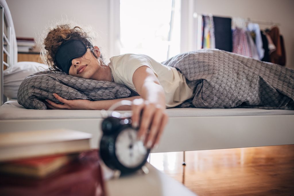 Woman in bed with sleep mask on holding the alarm clock going off
