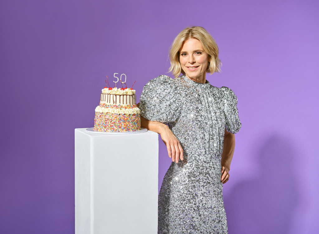 Emilia in silver dress leaning against plinth with cake on