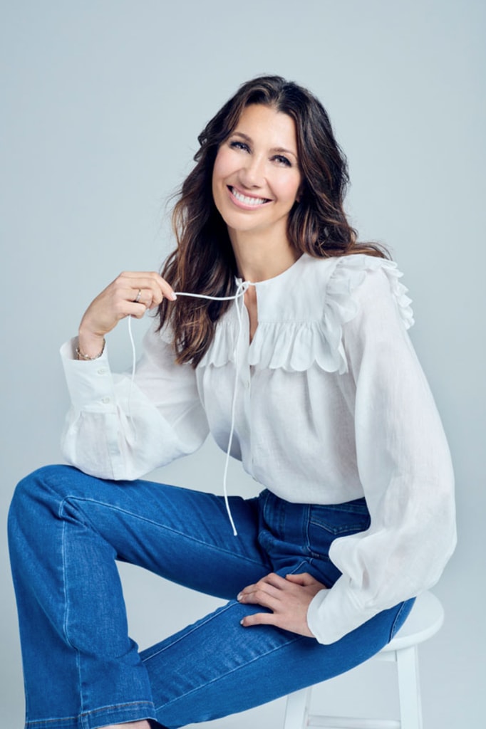 brunette woman in white top and blue jeans holding the string of her top and smiling 
