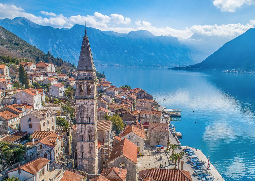 An aerial view of Perast, Montenegro, shows a historic stone bell tower rising above red-tiled roofs of buildings clustered along the bright blue Bay of Kotor. Majestic mountains frame the water in the background.