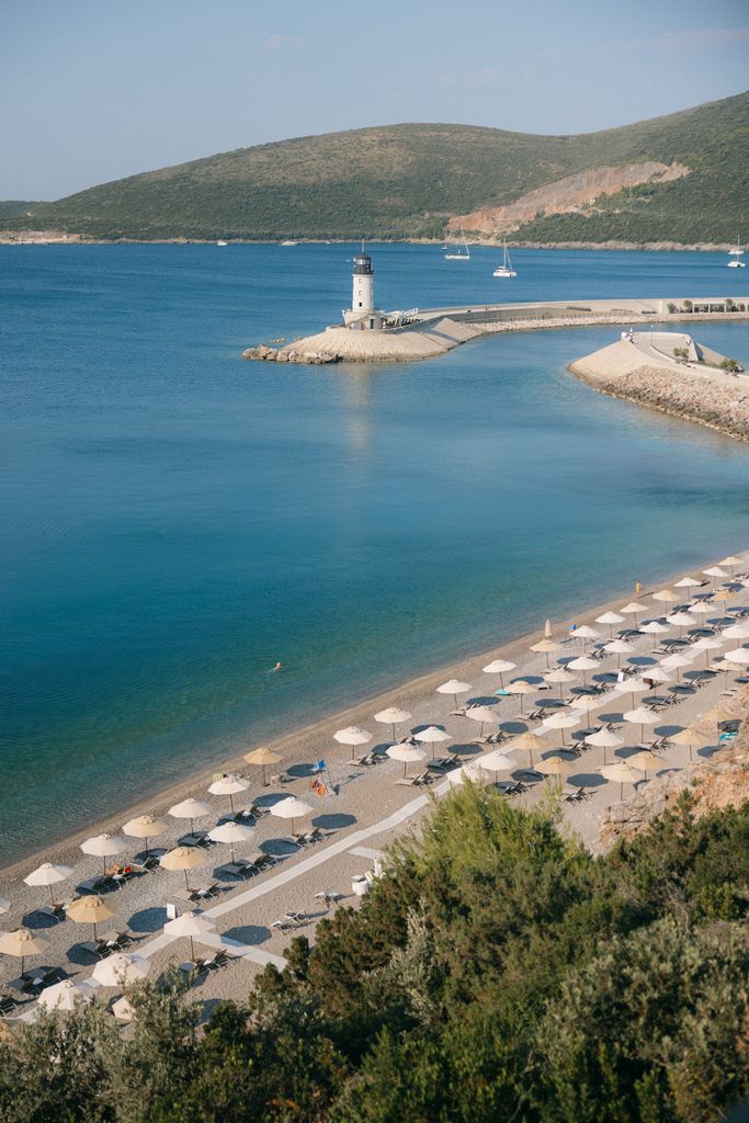 Lustica Bay, Montenegro, features a beach with umbrellas and loungers, a white lighthouse on a stone jetty, and calm blue water with distant boats, all set against green hills.