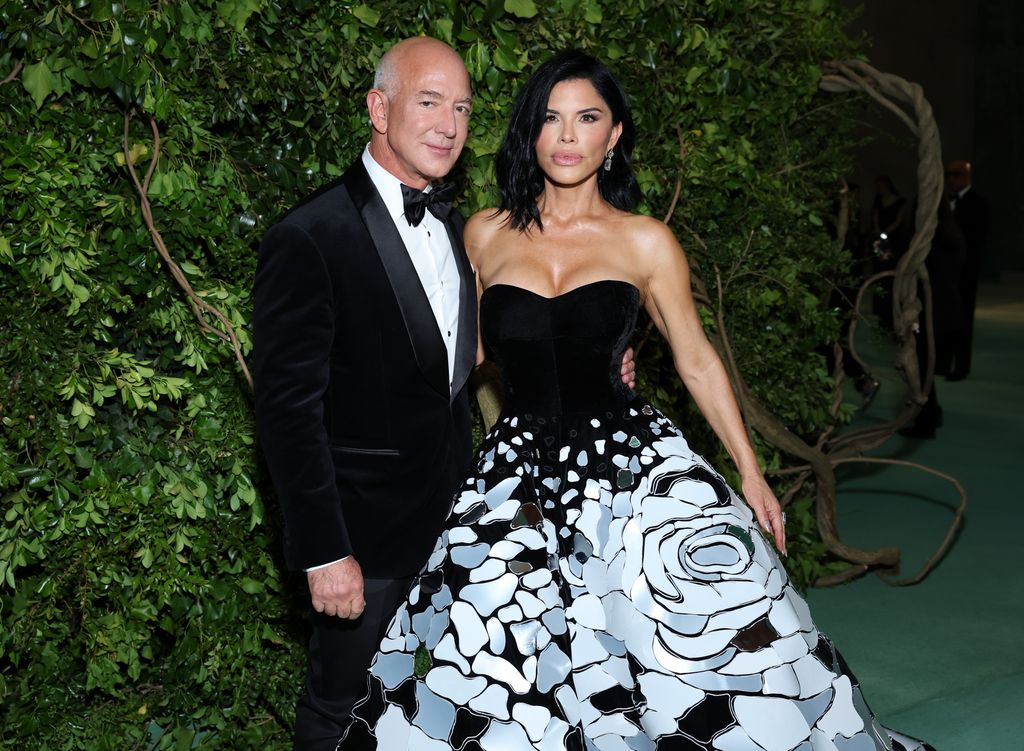 Jeff Bezos in a tuxedo and Lauren Sanchez in a black dress posing in front of green backdrop at the MET Gala