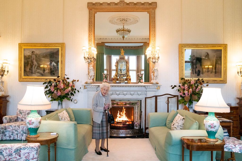 The Queen standing in a light room with green sofas, floral lamps and a fireplace in the centre
