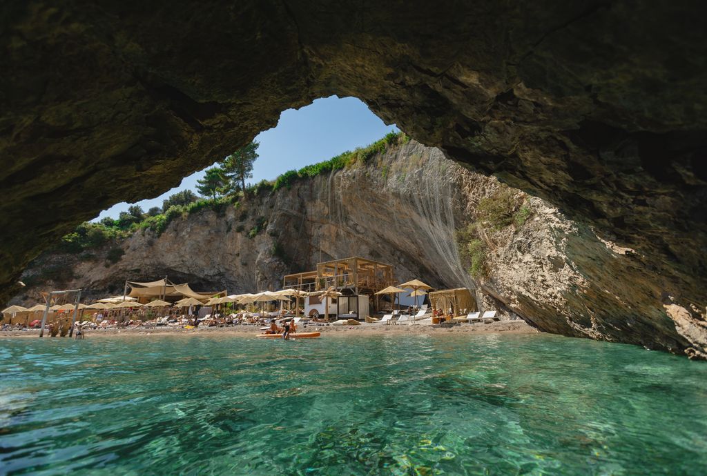 A view from inside a sea cave on a beach in Lustica Bay, Montenegro. The cave entrance frames a narrow beach with lounge chairs and umbrellas, and people relaxing by the turquoise water. 