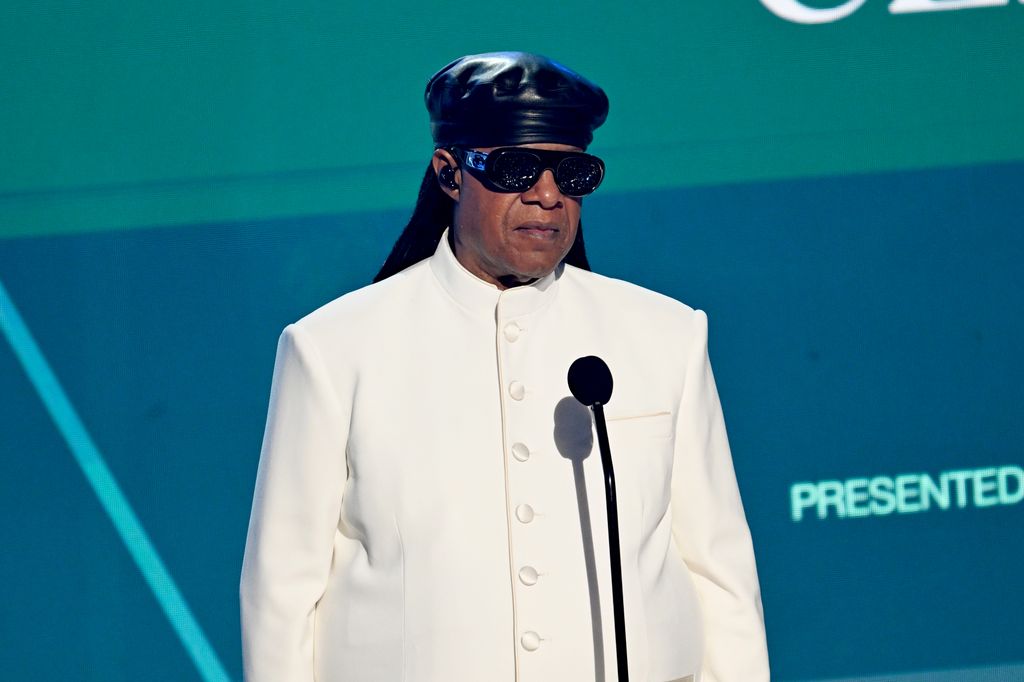 stevie wonder in white jacket and black hat on stage in front of microphone