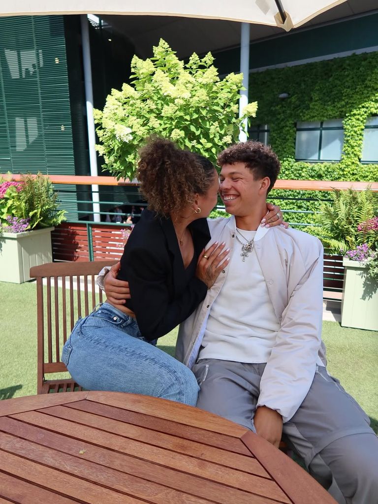 Trinity Rodman and Ben Shelton sitting at a wooden table at Wimbledon AELTC hugging and smiling at each other