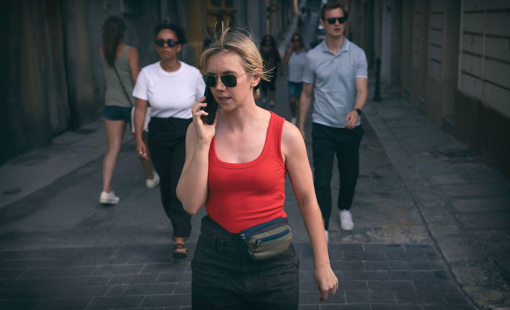 blonde woman on phone in red top walking down street, followed by woman and man in sunglasses