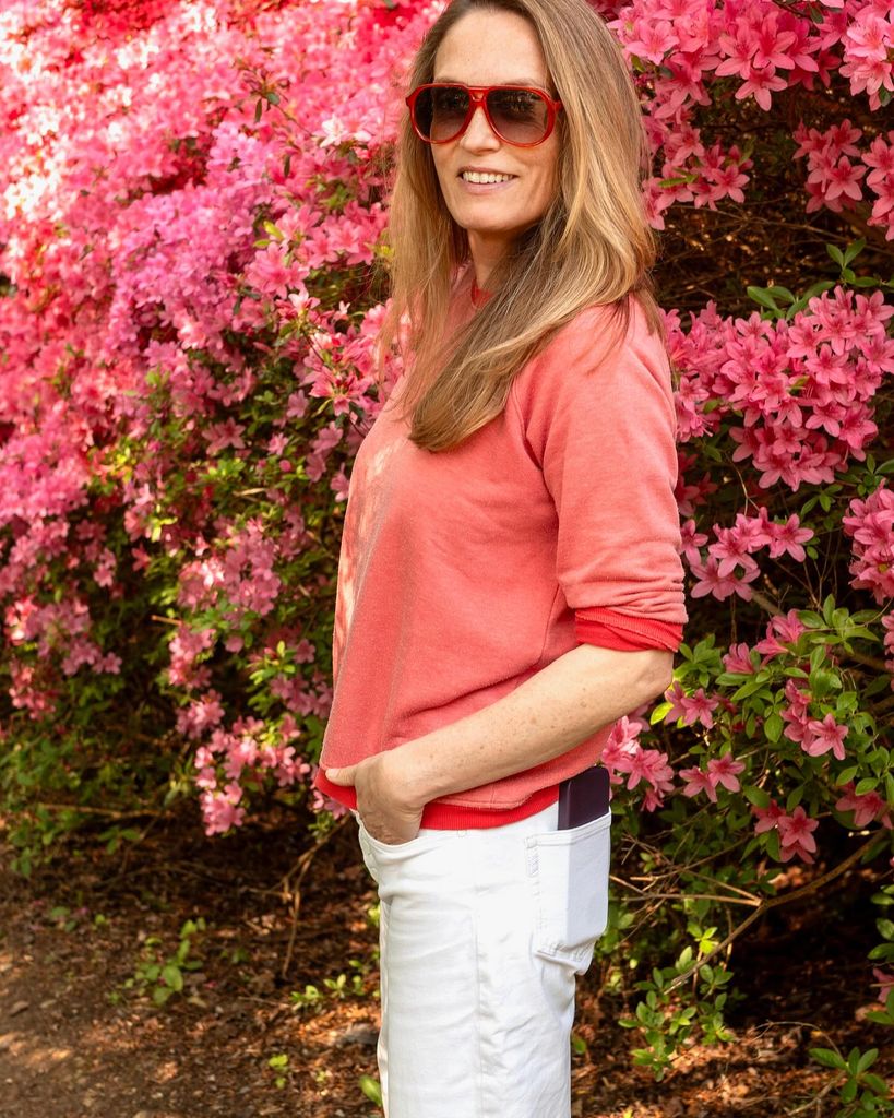 woman in pink top and white jeans smiling in front of a floral bush