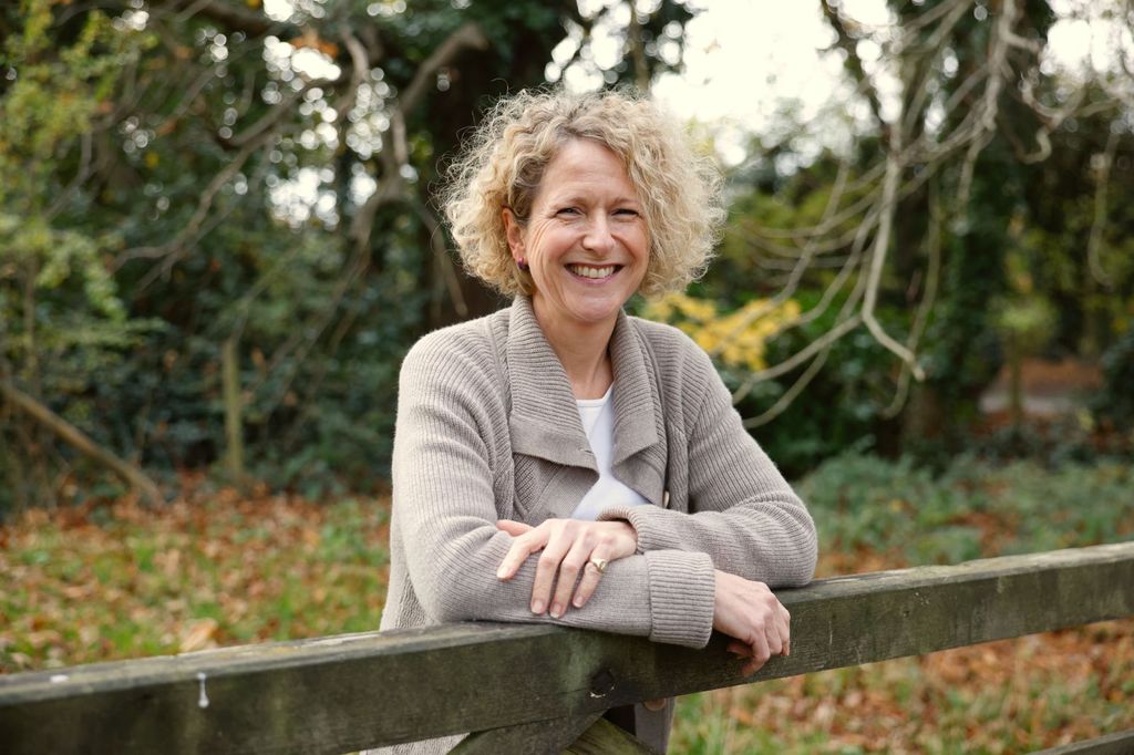 blonde woman with curly hair smiling and leaning on a gate