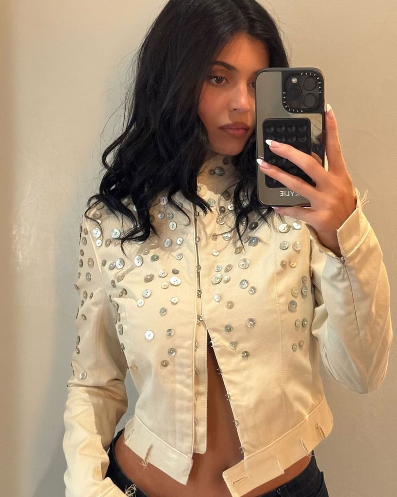 Kylie Jenner takes a mirror selfie, wearing a cream moto style jacket covered in silver buttons