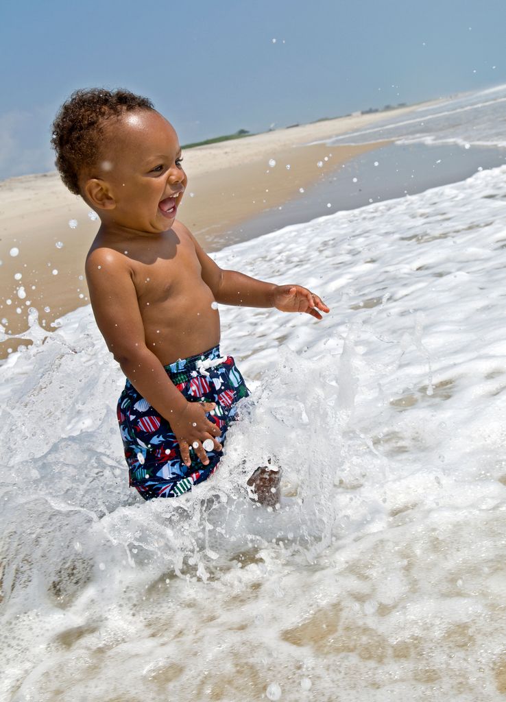 A baby boy standing on the beach with waves going past his lower body
