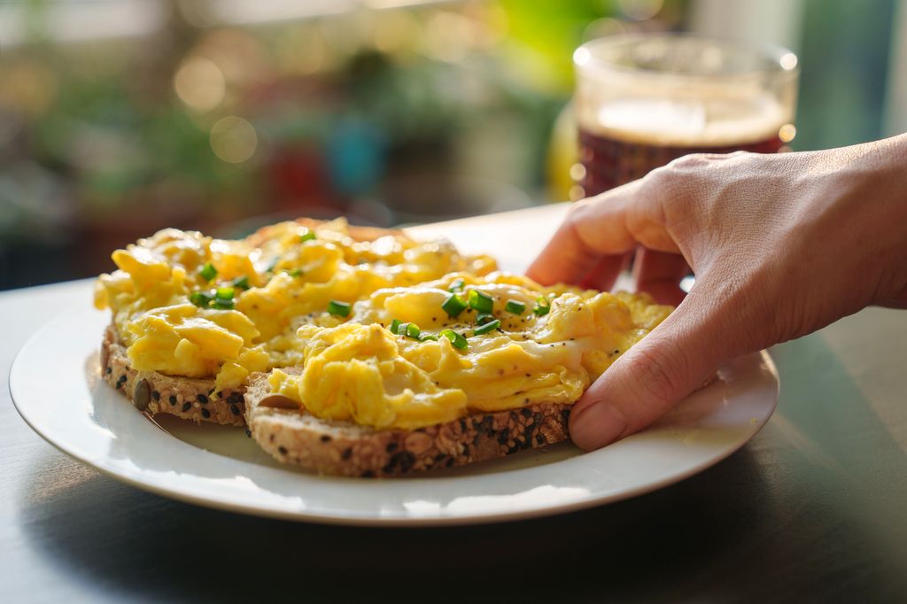 A cropped image of a woman's hand holding a piece of toasted bread with scrambled eggs on top, as she enjoys a healthy breakfast in the morning