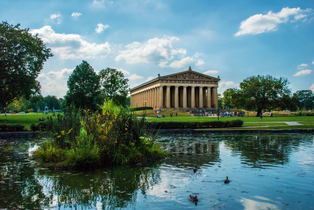 In late afternoon sunlight, the Parthenon can be seen against a bright blue sky, reflected in pond water at Centennial park in Nashville, Tennessee