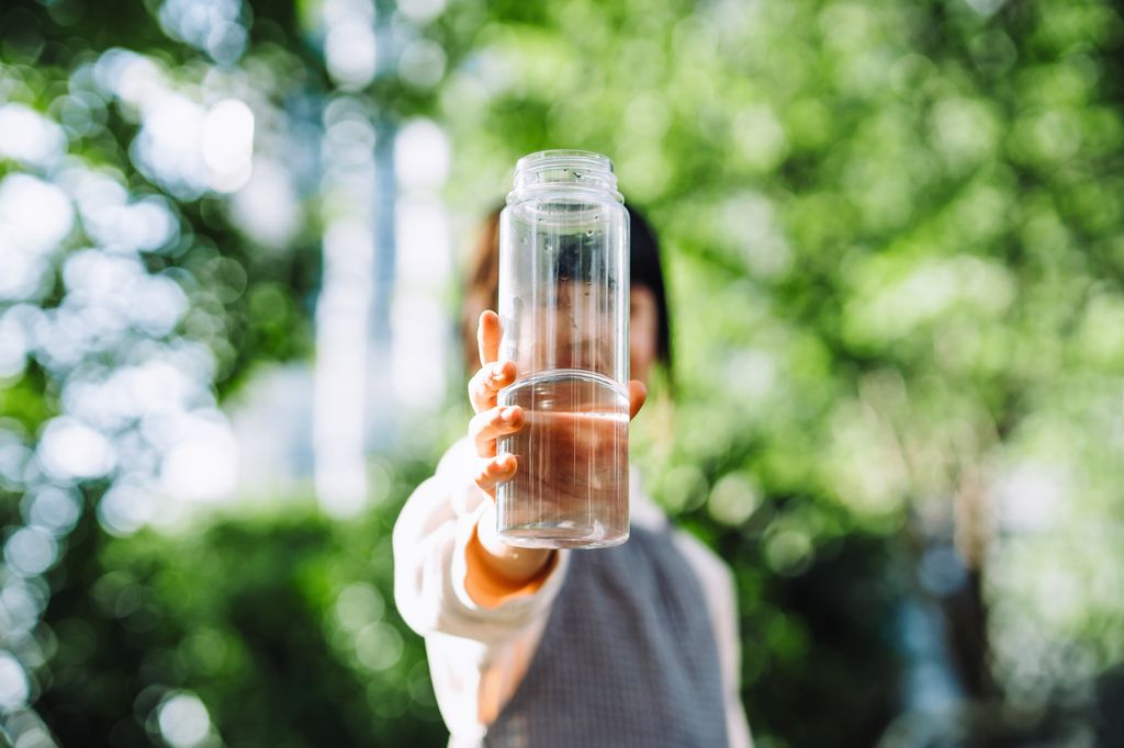 Portrait of Asian girl holding a reusable plastic bottle in front of her face, drinking water in park outdoors against sunlight with green plants. Staying hydrated. Healthy lifestyle. Environmental friendly, zero waste and sustainable lifestyle concept
