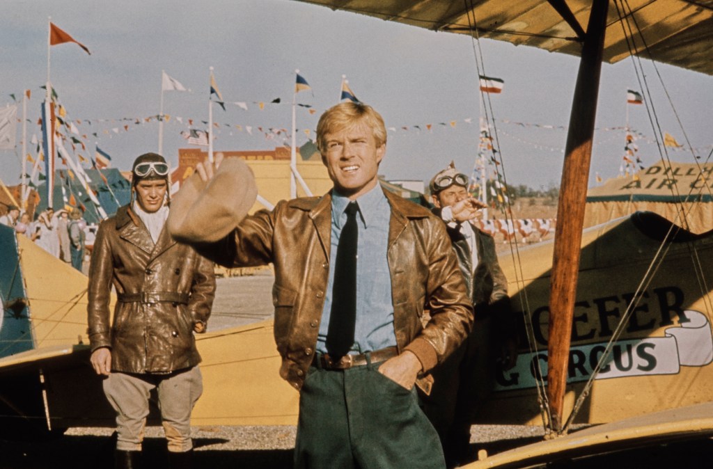 American actor Robert Redford, wearing a brown leather jacket over a blue shirt with a black tie, his cap in his right hand, with American actor Scott Newman, who wears a brown leather jacket, white scarf, an aviator's cap and flying goggles, a 'Dillhoefer's Flying Circus' biplane, bunting and flags in the background, in a publicity still from 'The Great Waldo Pepper', filmed in Elgin, Texas, 1975. The aviation drama, directed by George Roy Hill, starred Redford as Waldo Pepper. (Photo by Michael Ochs Archives/Getty Images)
