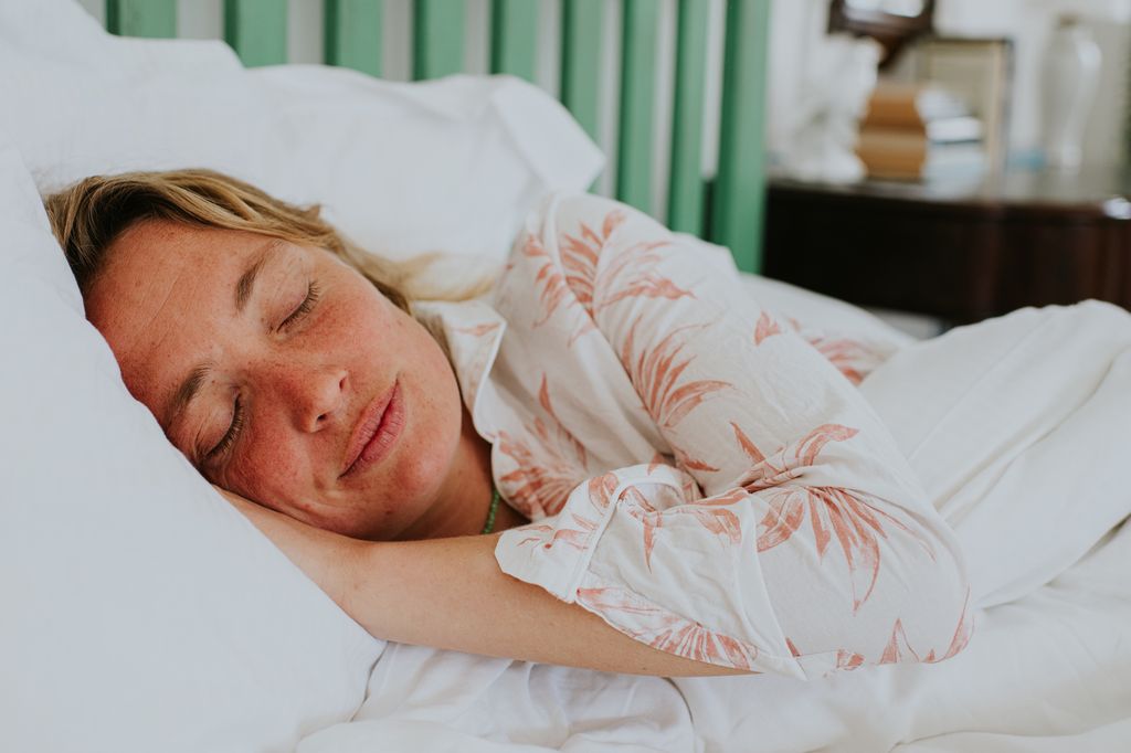 A young woman lies in a comfortable, soft double bed in a stylish airy bedroom. She looks utterly content and peaceful. She wears a pair of linen shorty pyjamas. Focus on her make-up free face.