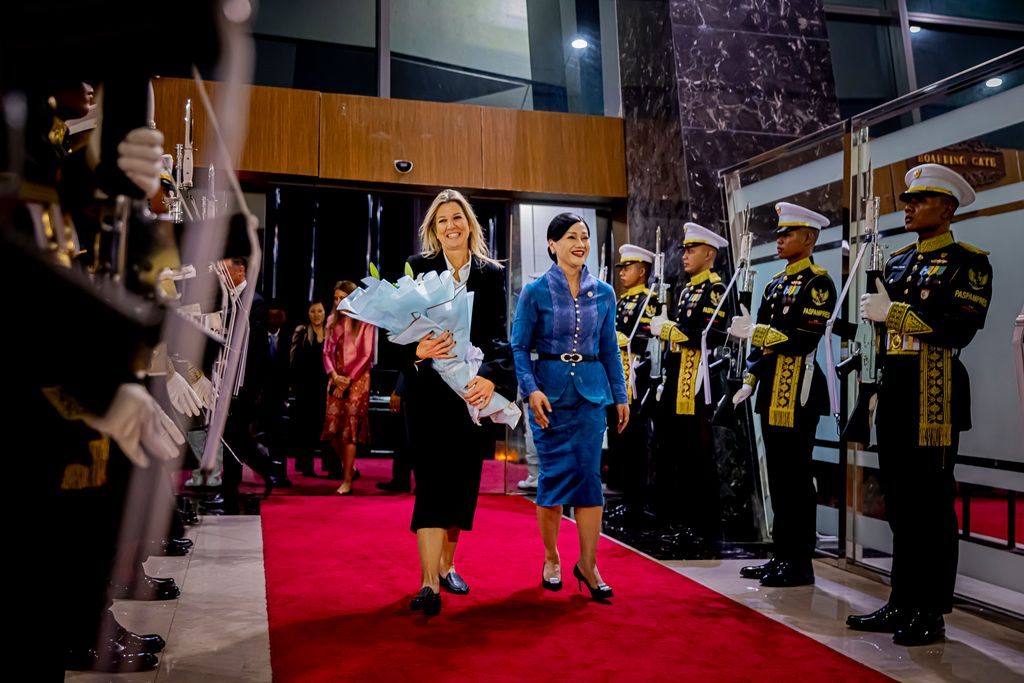 Queen Maxima holding flowers being greeted with military standing to attention each side of red carpet walkway
