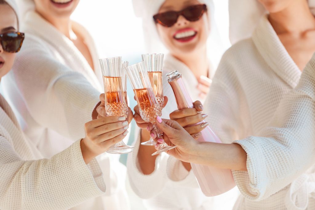 Group of cheerful young women wearing white bathrobes, towels on heads and sunglasses preparing for a bachelorette party. They toasting with champagne. Close up of champagne flute.