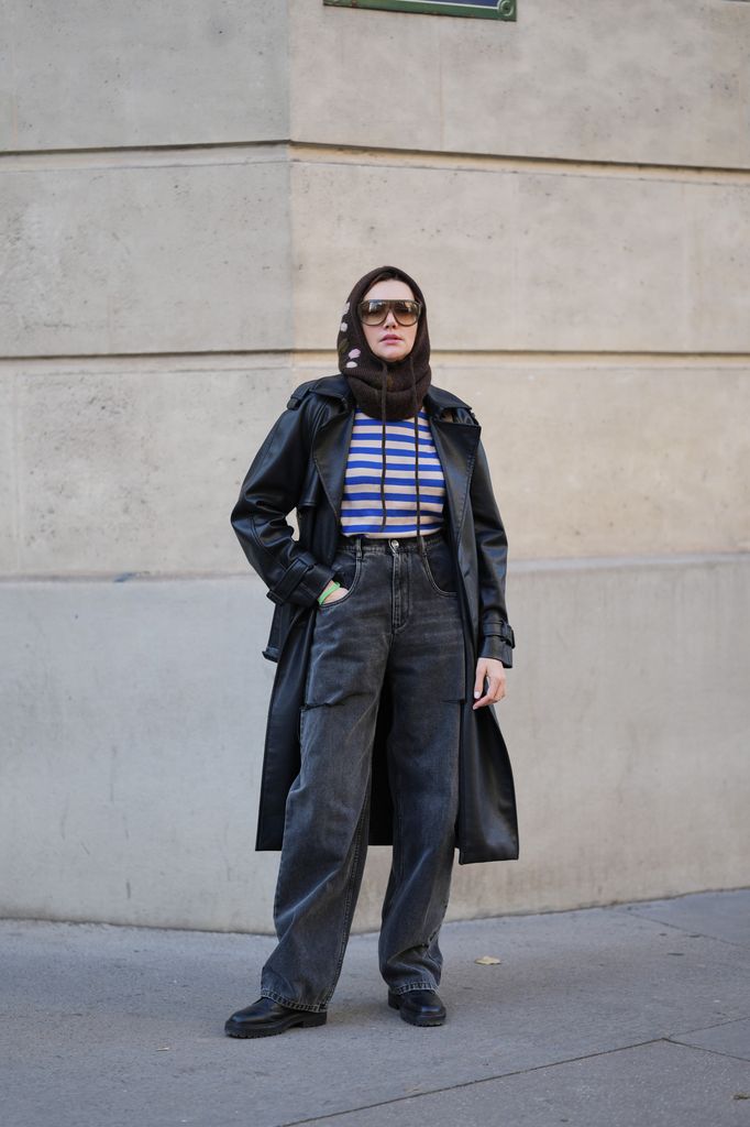 Olesya Senchenko wears a brown knit balaclava with small floral details by Zara,  oversized rectangular sunglasses by Jimmy Fairly, a blue and beige horizontal striped long sleeve top by LO NEEL, a black leather trench style jacket by H&M, high waist faded black wide leg jeans by Maison Margiela, black leather lace up shoes by Jimmy Choo.