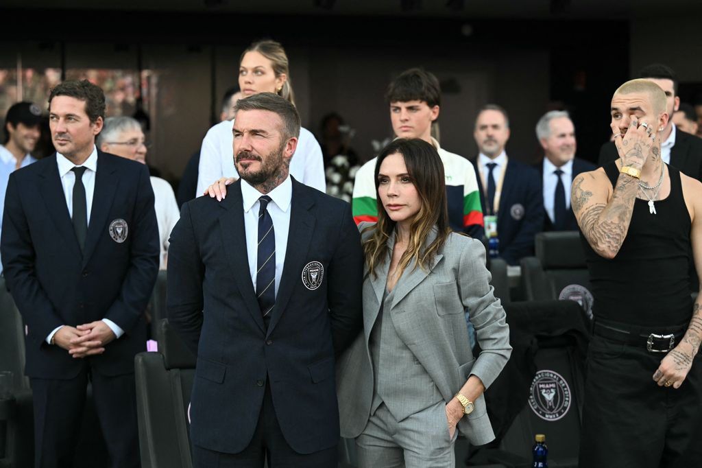 Victoria Beckham in a grey three-piece suit in group of people including david watching football