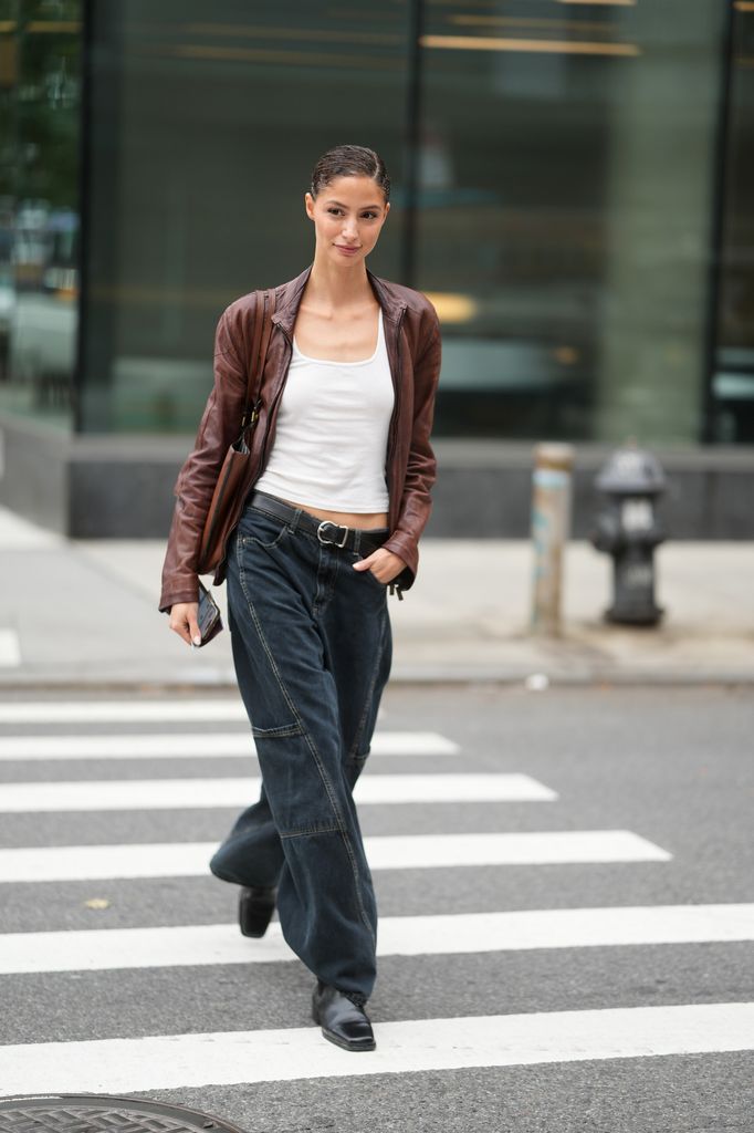 Camila Rodrigues is seen wearing a brown leather jacket, white tank top, black belt, oversized denim jeans, black boots and a brown shoulder bag outside of the Patbo show, during New York Fashion Week
